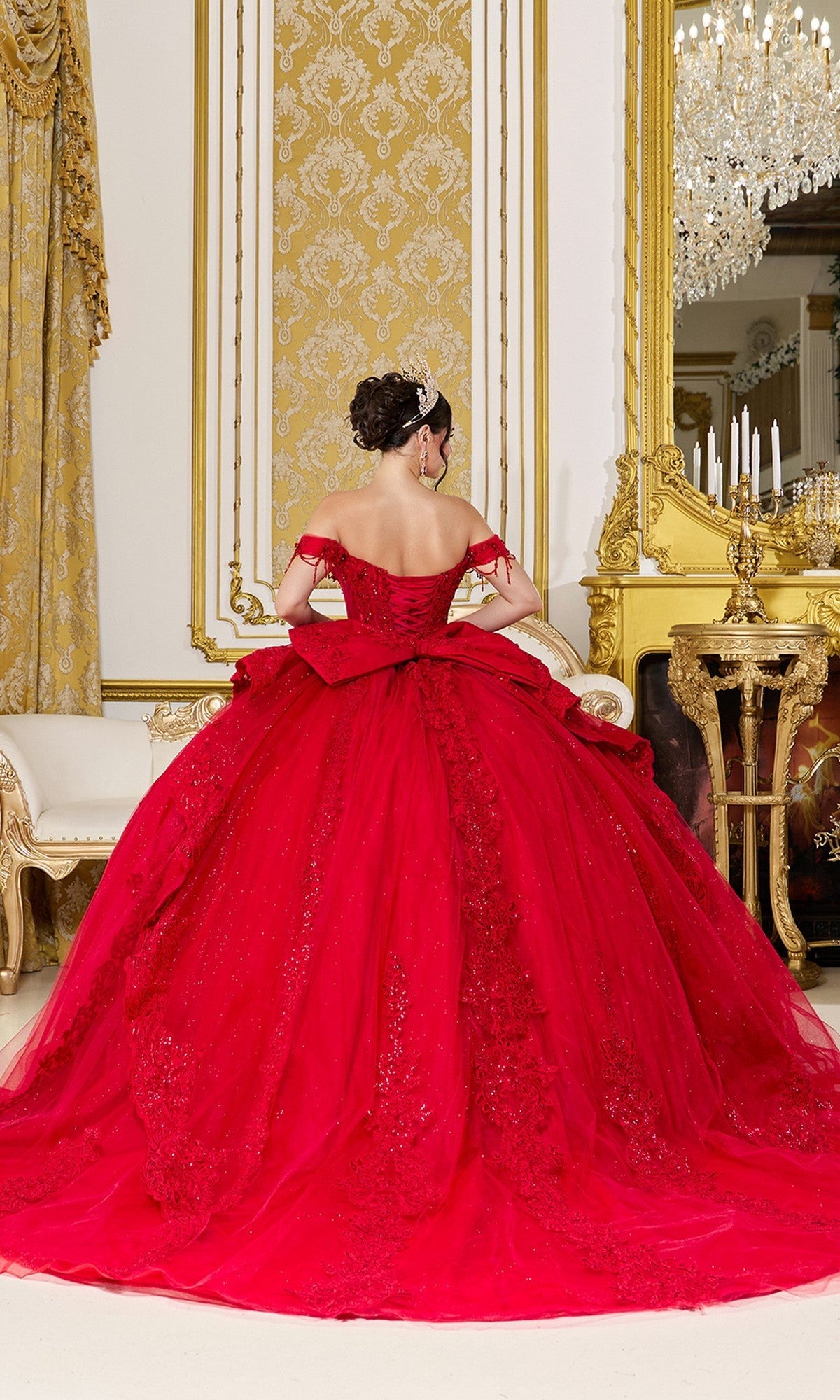 A woman wearing the Quinceanera Dress 5108 by Dancing Queen, featuring red glitter tulle and a corset bodice, sits on a cream and gold sofa in an ornate room with gold accents and a chandelier.